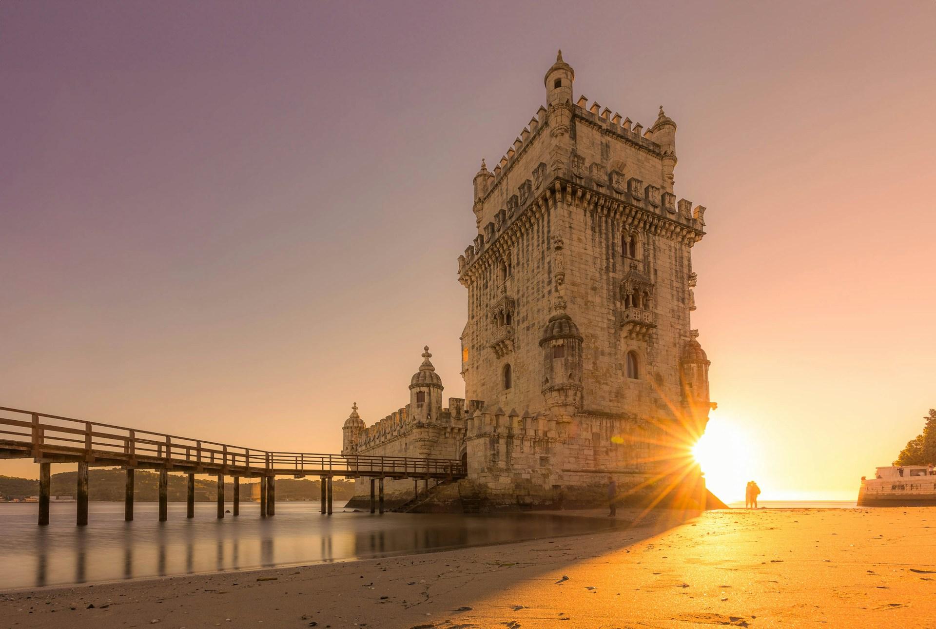 Belém Tower beside the Tagus River at sunset with two visitors on the shore in Lisbon, Portugal