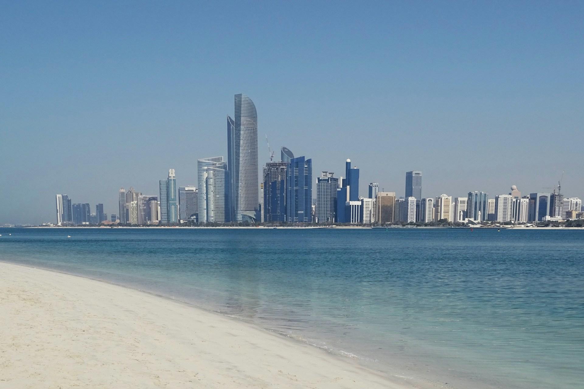 Abu Dhabi waterfront skyline viewed across blue water and a sandy beach in the United Arab Emirates